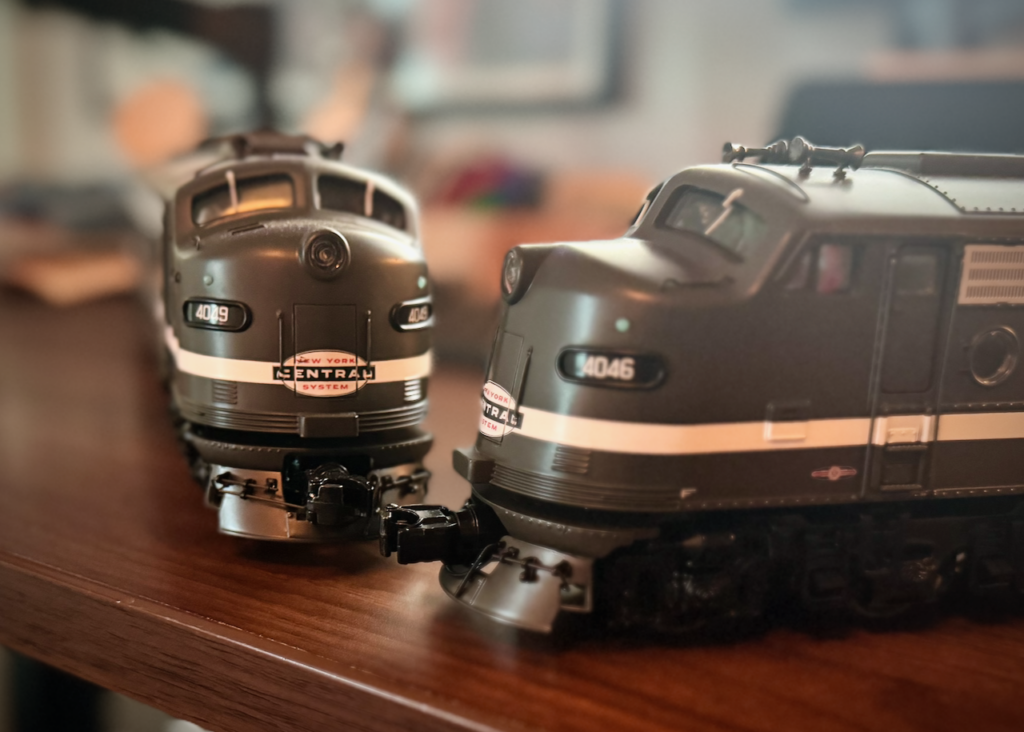 The image shows two model train locomotives placed on a wooden surface. They are painted in a dark green color with white stripes and have the "New York Central System" logo prominently displayed on their fronts. The locomotive on the left has the number "4049" and the one on the right has the number "4046." Both locomotives have a streamlined design typical of mid-20th century diesel engines. The background is slightly blurred, keeping the focus on the two model trains.