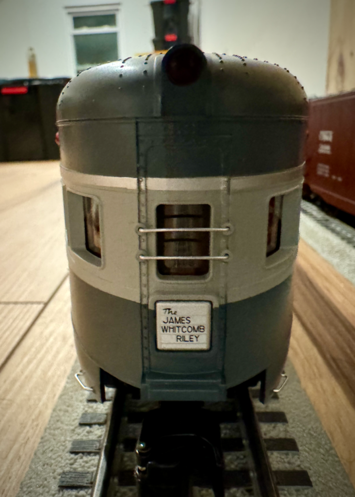 The image shows the rear end of a model train car, specifically a passenger car. The car is painted in a two-tone color scheme, with the top half being a darker color and the bottom half a lighter color. The windows have silver framing, and the back of the train car features a small sign that reads "The JAMES WHITCOMB RILEY." The model train is situated on a track, and the image captures the details of the car, including its rounded rear end and the texture of the paint.