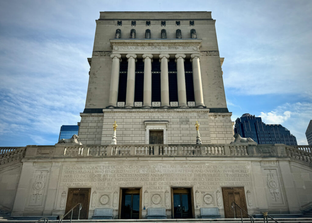 The image shows the north side of the Indiana World War Memorial, a monumental neoclassical building in Indianapolis. The structure is adorned with tall columns and inscriptions commemorating the valor and sacrifice of the United States military forces. The sky is clear, enhancing the grandeur of the memorial against the urban backdrop.