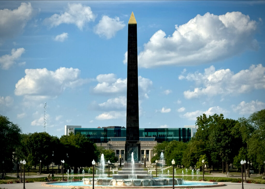 The image shows the Veterans Memorial Plaza in Indianapolis, featuring a tall, dark obelisk with a golden tip at its center. In the foreground, there is a circular fountain surrounded by neatly landscaped grounds. The Indianapolis Central Library, with its modern glass facade, is visible in the background under a clear, partly cloudy sky.