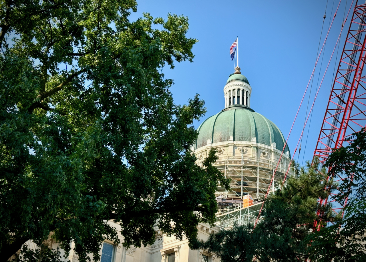 This photo captures the dome of the Indiana Statehouse, partially obscured by a large tree with lush green leaves in the foreground. The dome itself is an elegant structure with a patina-green color, characteristic of weathered copper. The dome is topped with a small cupola, and the U.S. flag and Indiana state flag are flying from the top. Scaffolding surrounds the base of the dome, indicating that some renovation or maintenance work is underway. To the right of the image, a red construction crane is visible, suggesting active work on or around the dome. The sky is clear and blue, providing a bright backdrop to the scene.