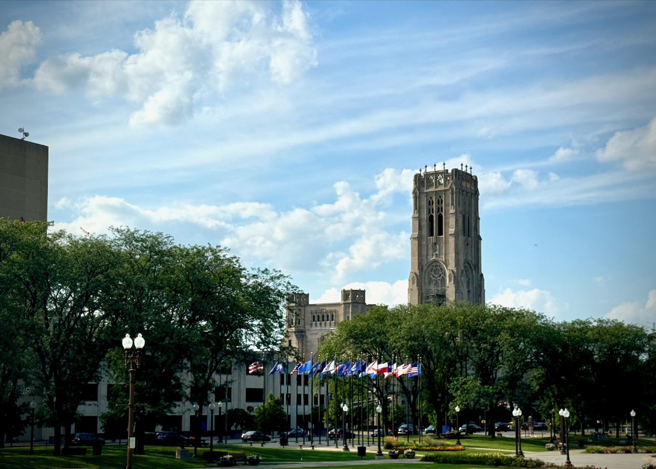 The image shows the tall, ornate tower of the Scottish Rite Cathedral in the background, with its Gothic architecture standing out against a partly cloudy sky. In the foreground, Veterans Memorial Plaza is visible, featuring a row of flags and green spaces surrounded by trees. The overall scene combines historical architecture with a tranquil urban park setting.