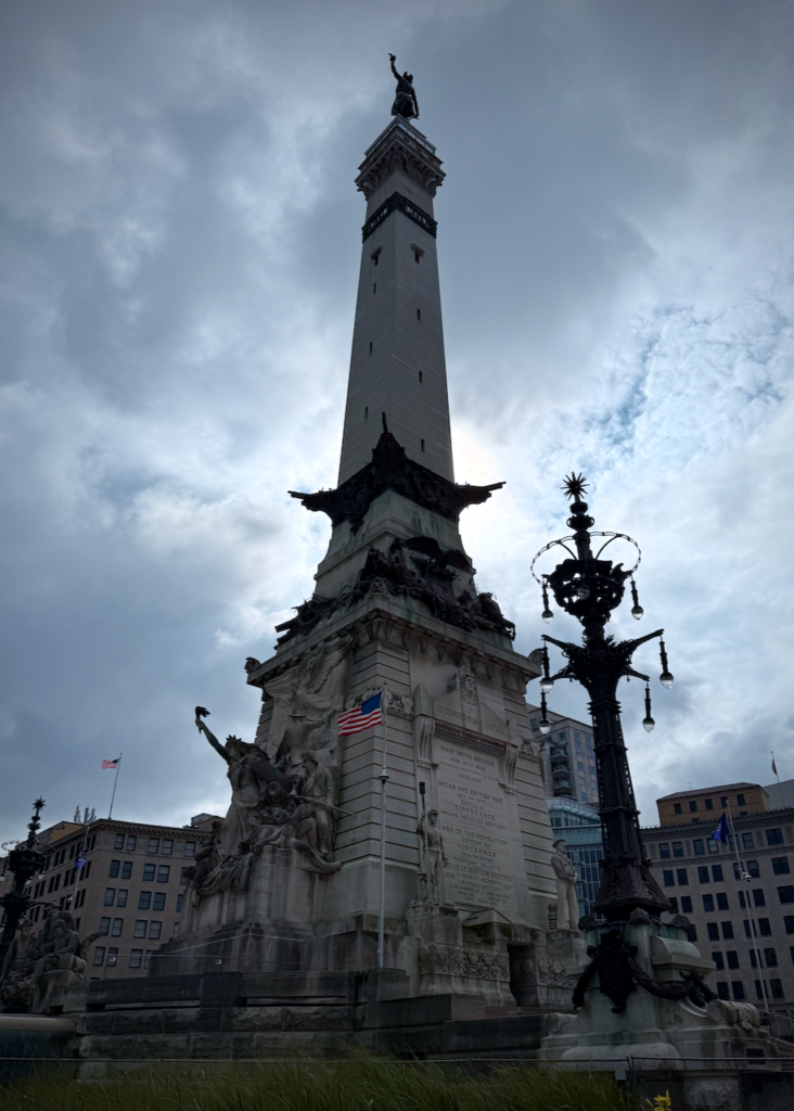 This is the Soldiers' and Sailors' Monument in downtown Indianapolis, set against a cloudy sky. The monument features intricate sculptures, including a group of figures representing military and allegorical themes. An American flag is mounted near the inscriptions on the monument's pedestal. The figure of Victory stands atop the monument, and an ornate streetlamp with a distinctive design is visible to the lower right.