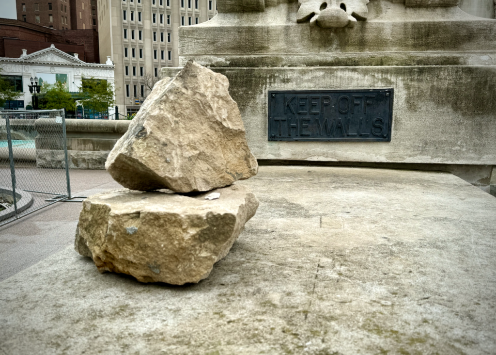 The image shows a close-up of a stone wall of the Soldiers' and Sailors' Monument in Indianpaolis. On the ledge are two large, stacked rocks. A metal plaque is affixed to the wall behind the rocks which reads, "KEEP OFF THE WALLS," as a warning. On the left you can see the Hilbert Circle Theatre and the AES Indiana headquarters.