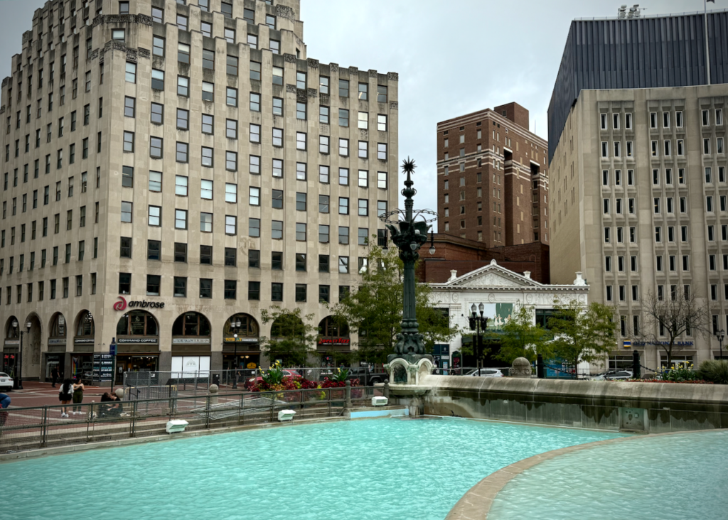 View of the southeast quadrant of Monument Circle, with a prominent water fountain pool in the foreground, part of the Soldiers' and Sailors' Monument. Across the street from the pools are several tall buildings, including an Art Deco high-rise on the left with a sign that says "ambrose" on it. To the right is the Hilbert Circle Theatre, and then the headquarter of AES Indiana. The tall brick building in the background is Symphony Center.