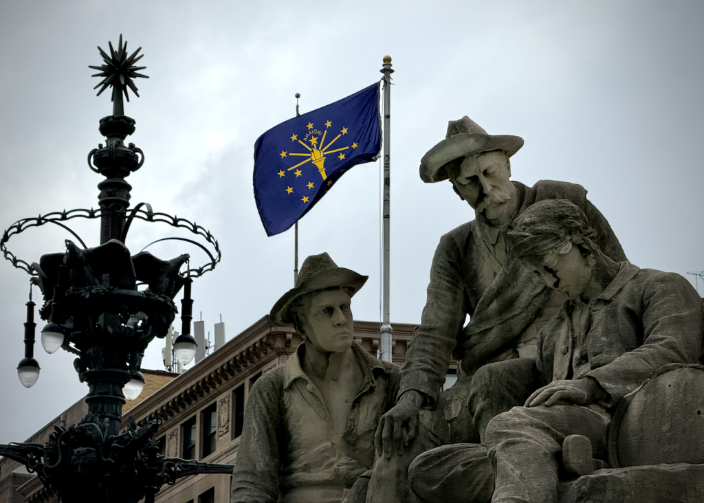 Close-up view of a limestone sculpture on the Soldiers' and Sailor's Monument in Indianapolis call The Fallen Soldier. One figure it slumped over while the others look at him with concern. Behind them is the Indiana state flag, with it's torch and stars emblem. A decorative streetlamp with a wrought iron design is also visible in the foreground.