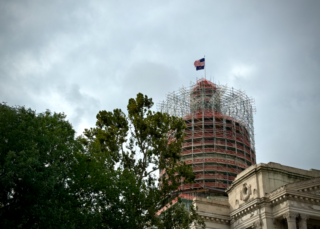 The image shows the dome of the Indiana Statehouse surrounded by scaffolding, indicating restoration work. Atop the dome flies the U.S. and Indiana flags. Green trees are visible in the foreground.