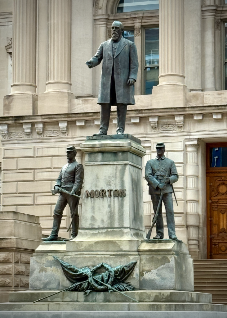 The image shows a statue of Oliver P. Morton, positioned in front of the Indiana Statehouse. Morton, a prominent figure in Indiana's Civil War history, stands at the top of the monument, with two soldiers depicted on either side at the base. The soldiers, dressed in Civil War uniforms, representing the Union, reflecting Morton's role during the war. The pedestal bears Morton's name, with a decorative laurel wreath and flags at the bottom.