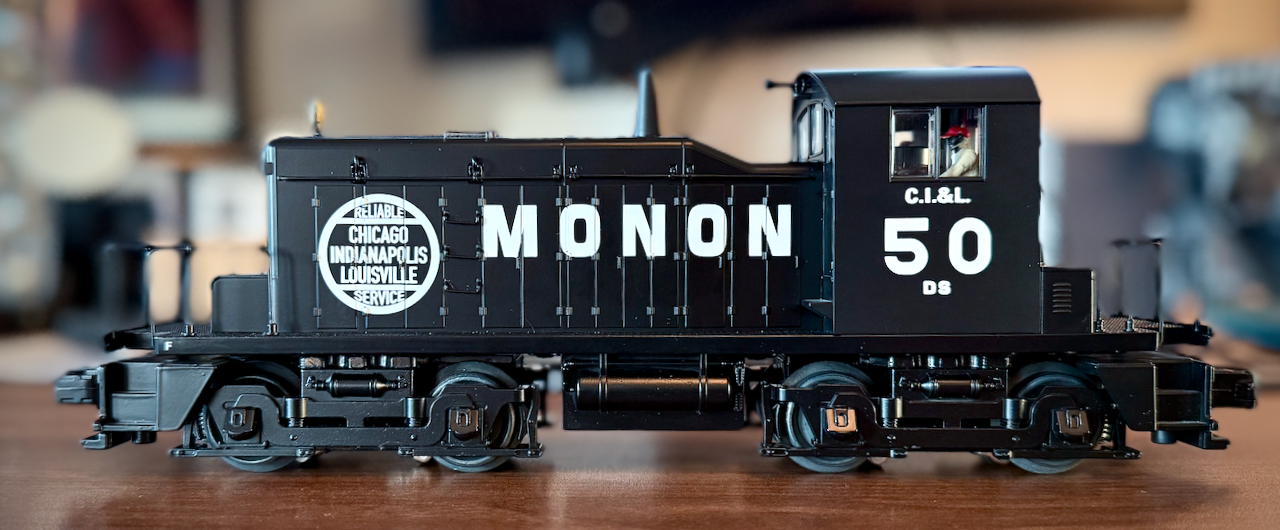 This image shows a detailed model of the Monon DS-50 locomotive. The black engine features bold white lettering reading "MONON" on the side, along with a circular emblem that says, "Reliable Chicago Indianapolis Louisville Service." The number "50" and is visible on the cab. The model is set on a table, with a softly blurred background, highlighting the craftsmanship of this vintage railroad replica.