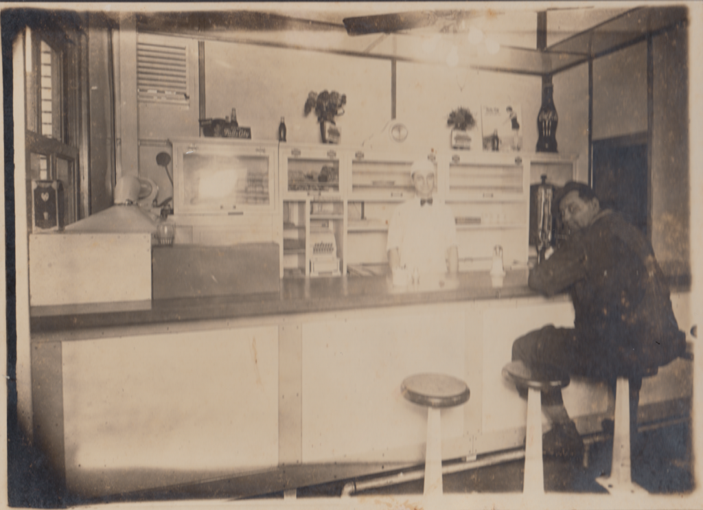 A vintage interior of a 1920s restaurant, featuring a counter with stools. A worker in a white uniform and bow tie stands behind the counter, while a man in dark clothing sits on one of the stools, leaning slightly. The counter displays various containers and items, with shelves behind holding decor and products.