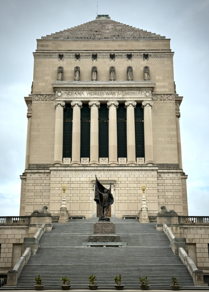 The image shows the Indiana World War Memorial, a large, rectangular monument with neoclassical architectural elements. Its inscription reads "Indiana World War Memorial" above a row of statues and columns. In front of the monument, a staircase leads up to a bronze sculpture titled Pro Patria, which depicts a soldier holding a flag, flanked by two stone sphinxes.