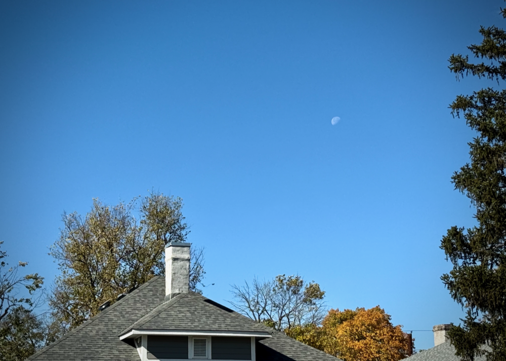 This image shows a house with a gray, shingled roof and a prominent white chimney in the foreground. Behind the house, there are trees with some fall foliage, including shades of yellow and orange. The sky is clear blue with a visible daytime moon in the upper right portion of the image. The scene feels calm, with a hint of seasonal change.