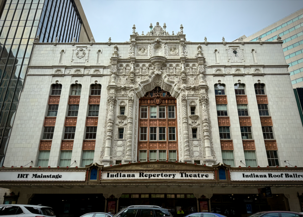 The image shows the Indiana Repertory Theatre, a historic building with an ornately detailed facade made of white terracotta. Marquees display the names "IRT Mainstage" (left) and "Indiana Roof Ballroom" (right).