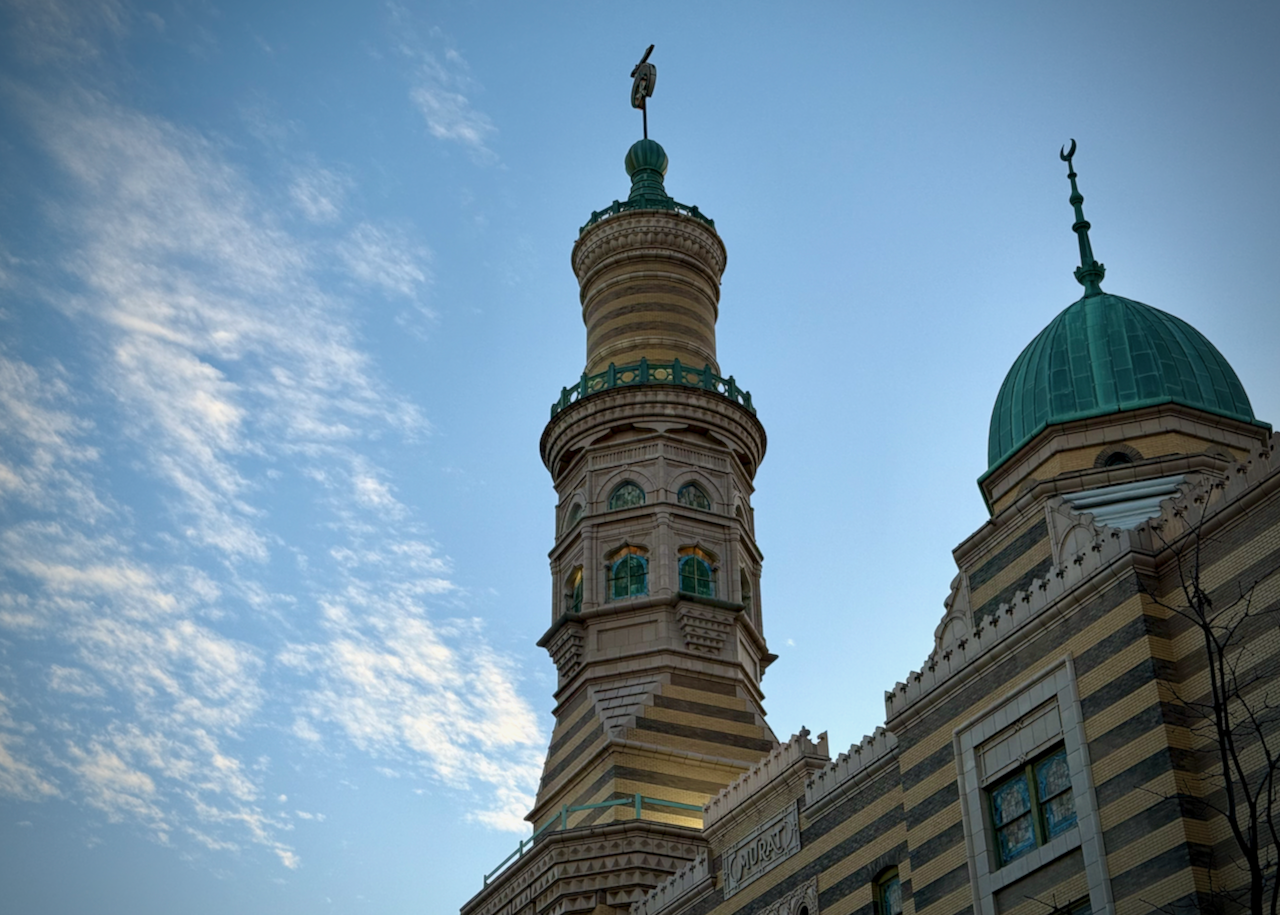 This image shows the iconic Murat Shrine, now known as the Old National Centre in downtown Indianapolis. The focus is on its Moorish Revival architectural elements, including a tall, striped minaret with intricate details, green accents, and a crescent-topped dome on the right. The sky provides a serene backdrop with scattered clouds, enhancing the building’s striking features.