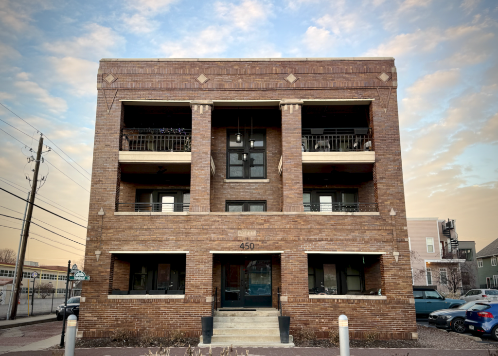 A brick apartment building called The Aviary, located in downtown Indianapolis along the Cultural Trail. The building features a symmetrical design with central double doors, two balconies, and decorative brickwork, under a sky with soft clouds at sunset.